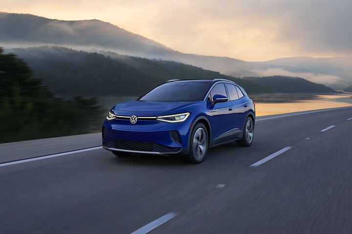 An ID.4 in Dusk Blue Metallic drives along a two-lane highway with mountains in the background.