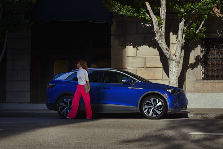A young family loads a bag into the cargo area of an ID.4, shown in Dusk Blue Metallic parked in a residential driveway.