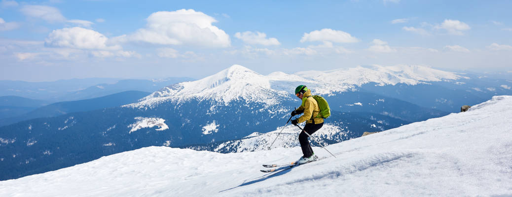Sportsman skier in helmet and goggles with backpack riding down steep snowy slope on copy space background of blue sky and beautiful mountain landscape. Winter holidays, extreme sport concept.