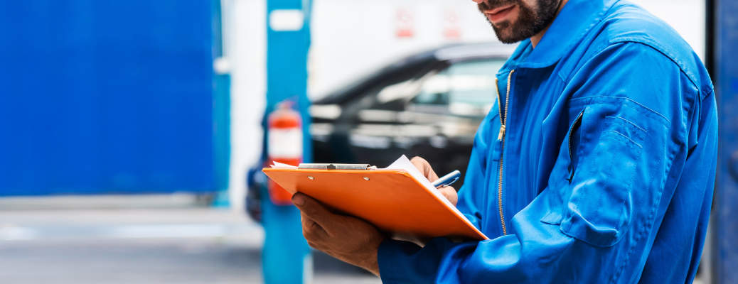Crop image of mechanic in blue work wear uniform checks the vehicle maintenance checklist with blur lifted car in the background. Automobile repairing service, Professional occupation.