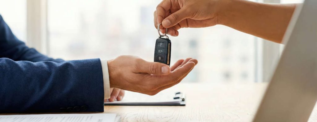 A man receiving a bad credit auto loan in Edmonton. A detailed shot of hands exchanging a car key between a salesperson and a client at an office desk with a laptop, reflecting a successful transaction. 