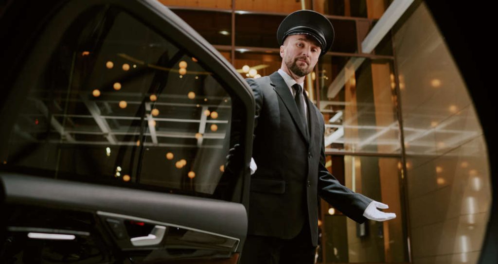 Caucasian middle aged man wearing uniform and cap opening car door with gloved hand, standing in modern building entrance, looking at camera, providing professional chauffeur service, representing what one might expect from the best valet service in the Design District