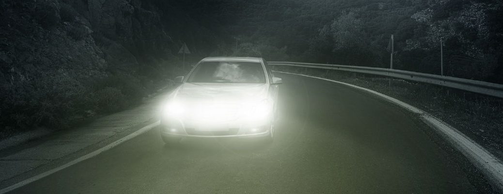 A photo illustration of a car driving on a dark road at night with its lights on.