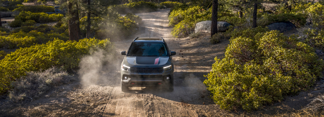 A 2025 Jeep Compass is shown driving on a dusty, off-road trail through a wooded area.