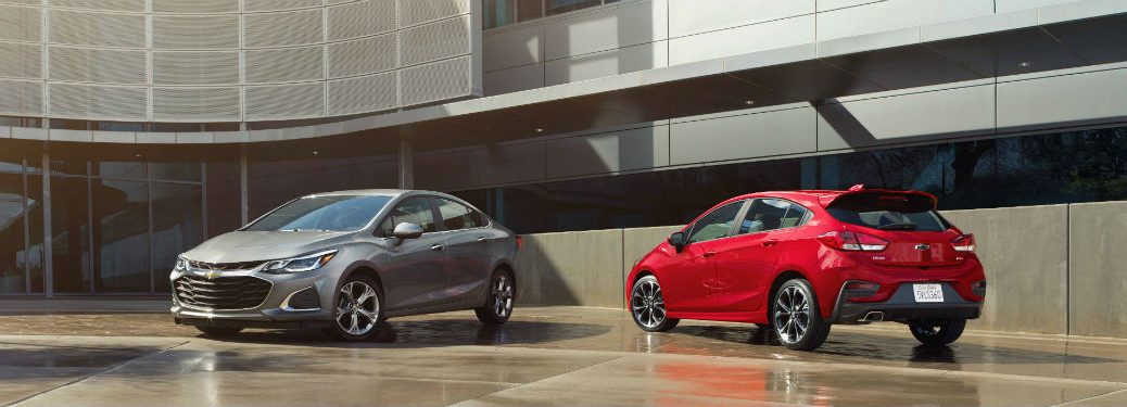 Driver side exterior view of a gray 2019 Chevy Cruze sedan on the left and a passenger side exterior view of a red 2019 Chevy Cruze Hatchback on the right