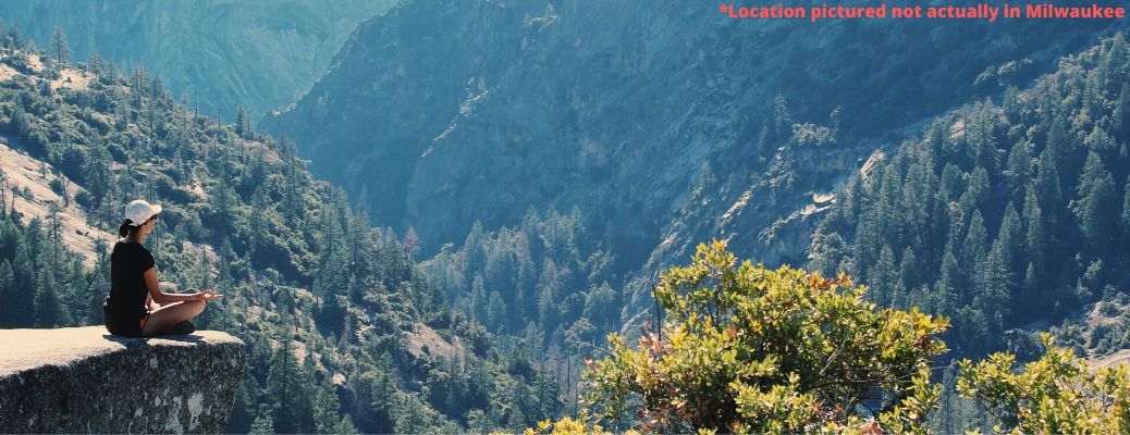 A woman meditates in a scenic mountain vista. Important disclaimer reads, "Location pictured not actually in Milwaukee"