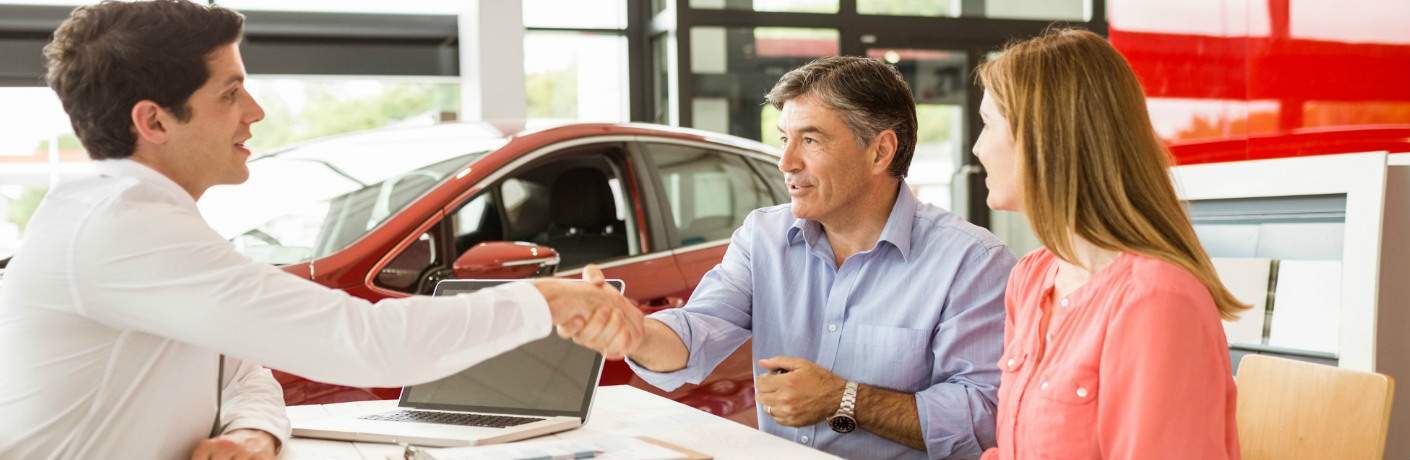 car salesman shaking hand of man and woman in dealership