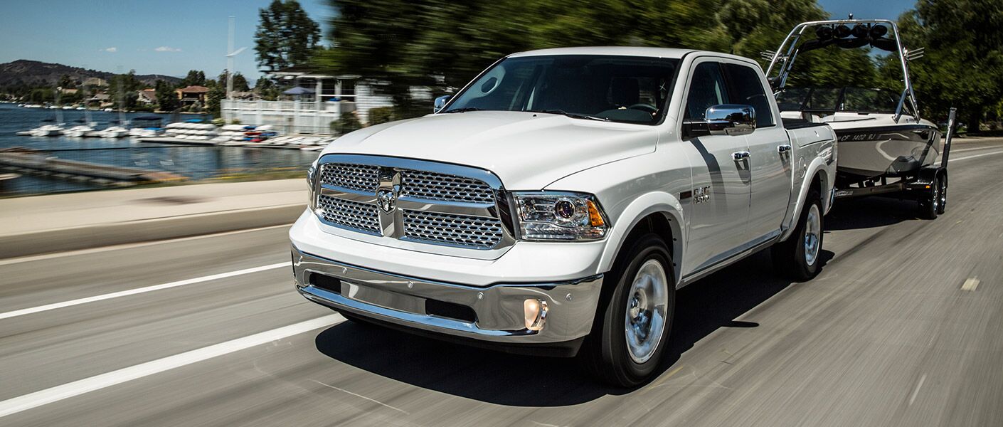 A white 2017 RAM 1500 truck tows a boat down a highway alongside a harbor.