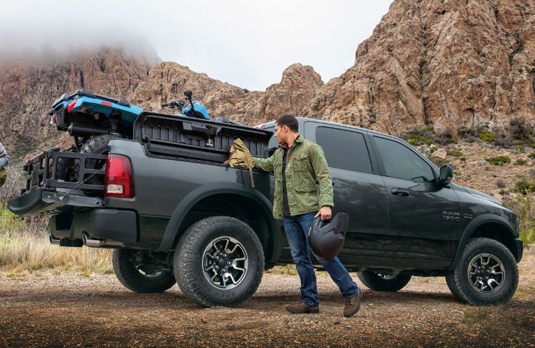 A man uses a cloth to wipe down the side of the bed of his pick-up truck, parked amidst craggy hills out in the desert. A recreational vehicle sits in the bed.