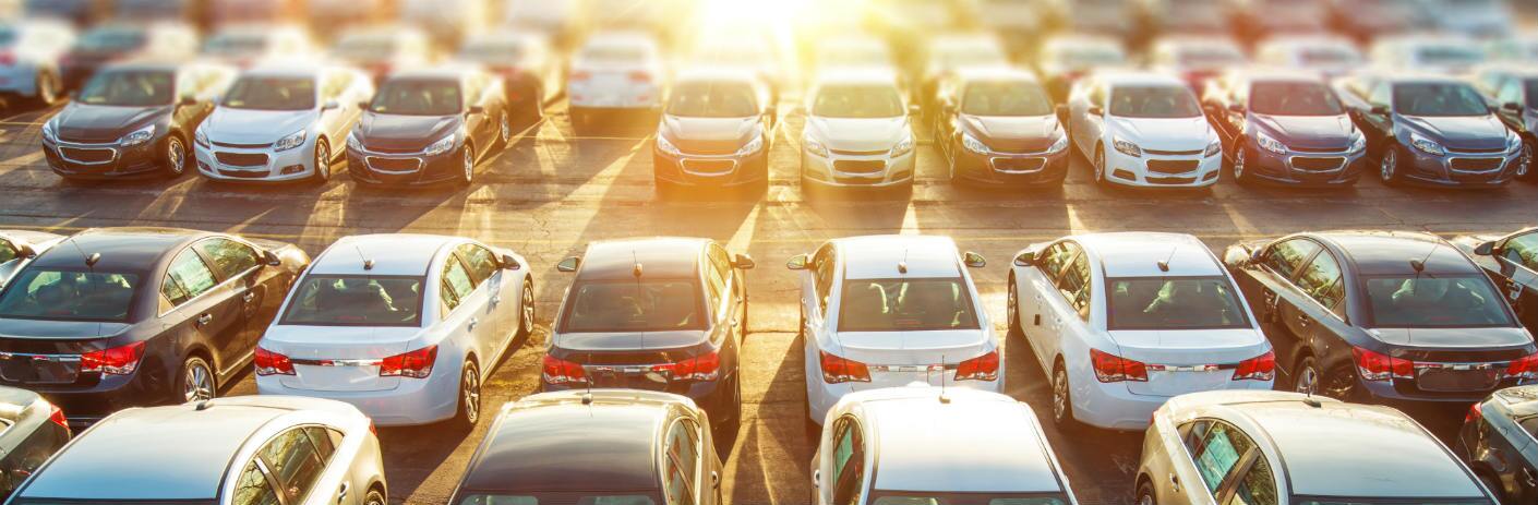 A multitude of cars are parked on a lot and seen from a raised angle. Bright sunlight blasts over them from the distance.