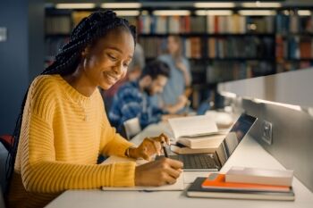 a black female college student researches in the library on her laptop