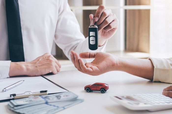 one person hands car keys to another over a desk with paperwork and money on it