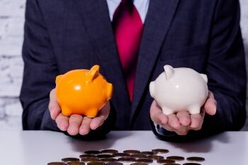 photo of a man in a suit holding an orange piggy bank in one hand and a white one in the other