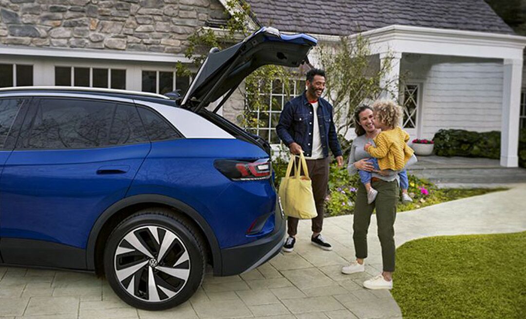 A young family loads a bag into the cargo area of an ID.4, shown in Dusk Blue Metallic parked in a residential home’s driveway.