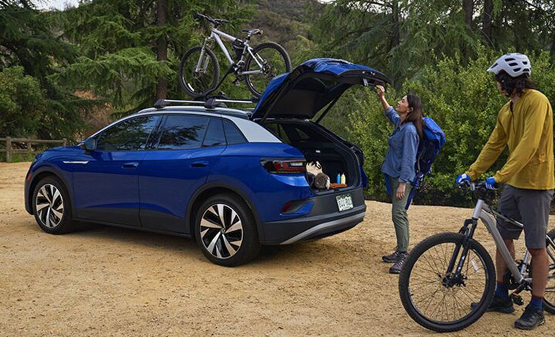 A man and woman load mountain bicycles onto an ID.4 in Dusk Blue Metallic with cross bars and a bike attachment that’s parked in a natural, outdoor setting.