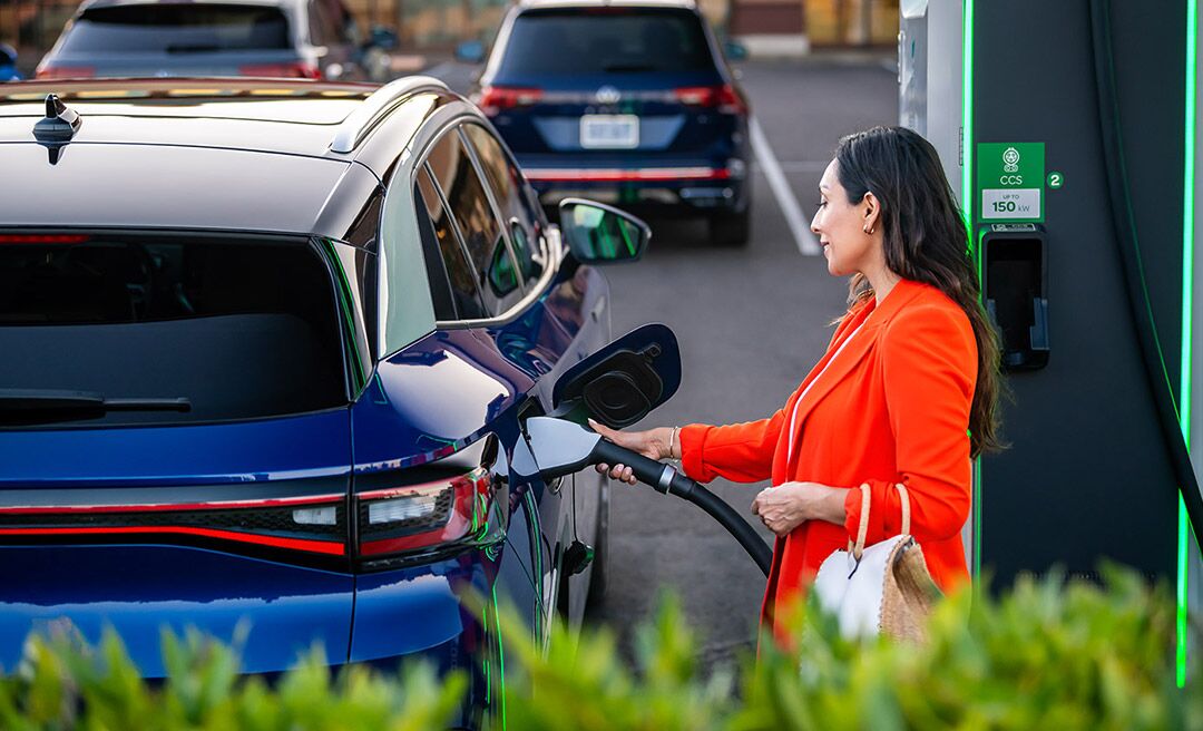 A well-dressed woman, parked at charging station stands on the passenger side of an ID.4 in Dusk Blue Metallic as she plugs in the car.