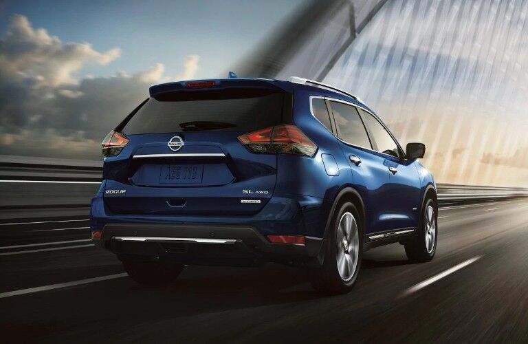 Rear passenger angle of a blue 2019 Nissan Rogue driving on a bridge towards a sunset