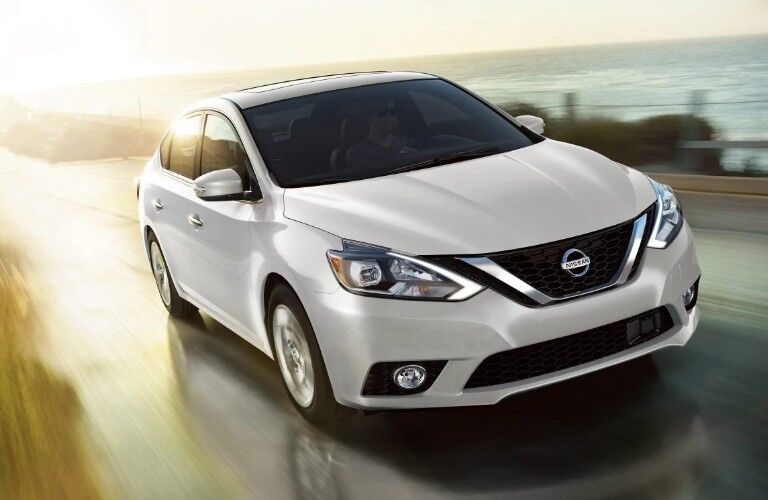 Front passenger angle of a white 2019 Nissan Sentra driving by the coast with a sunrise in the background