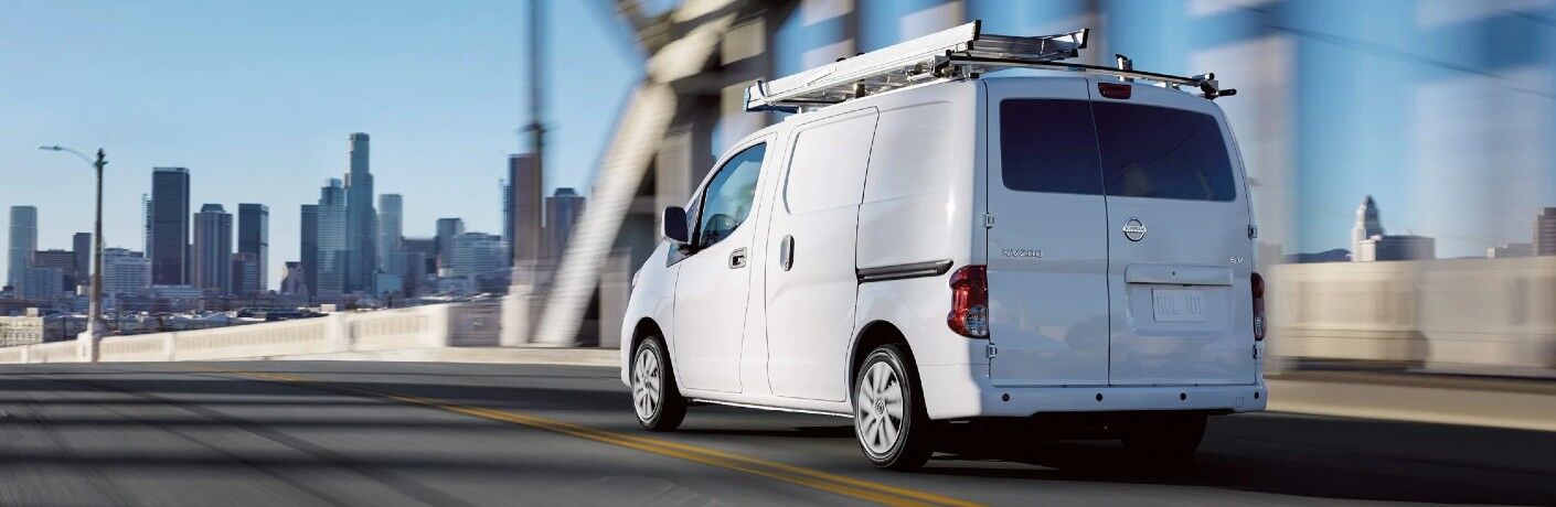 Rear driver angle of a white 2020 Nissan NV200 Compact Cargo driving towards a city