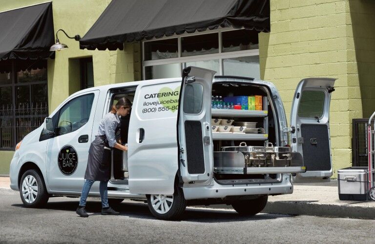 Rear driver angle of a white 2020 Nissan NV200 Compact Cargo with its doors open and an employee getting supplies out of it
