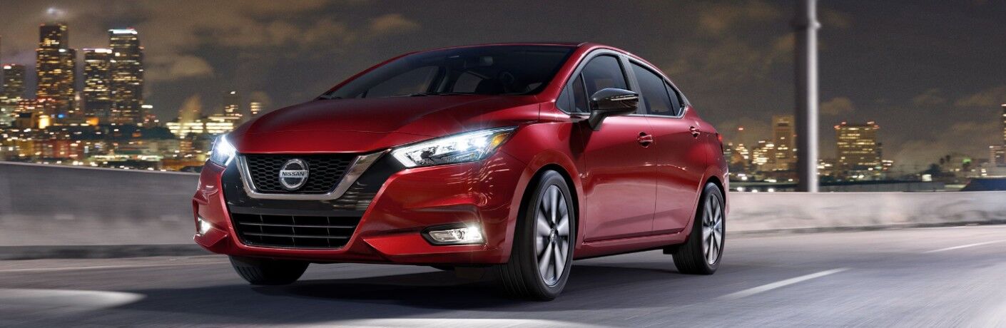 Front driver angle of a red 2020 Nissan Versa sedan driving down a road with a city in the background