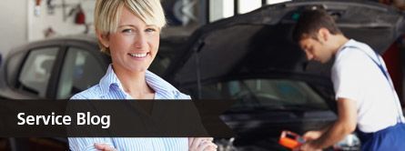 service blog text with woman looking at camera as mechanic works on her car behind her