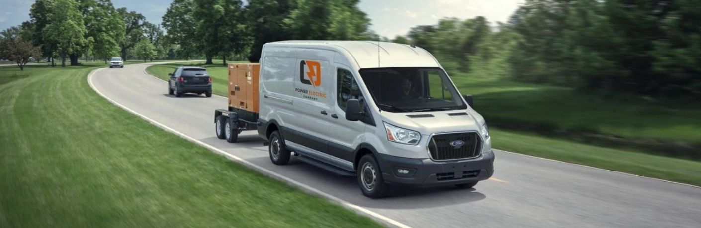 Front view of a white 2022 Ford Transit Cargo Van towing a n electric generator on a road surrounded by green grass and trees