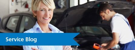 Woman smiling at camera with mechanic in background with "service blog" text on red banner