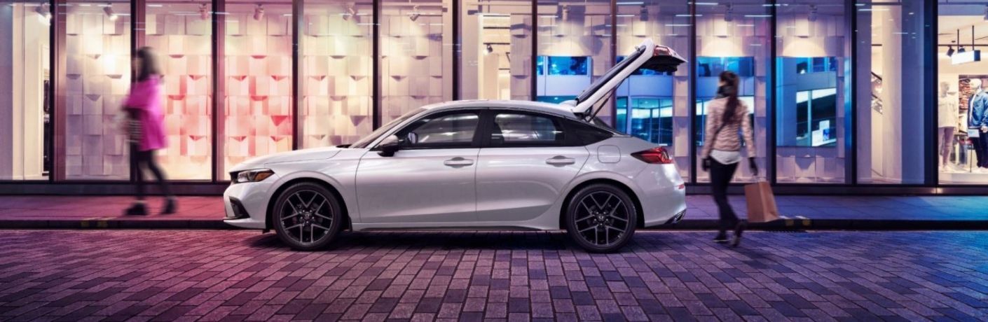 A woman carrying a shopping bag approaching the open trunk of a white 2022 Honda Civic Hatchback parked on a paved street in front of glass shops at night