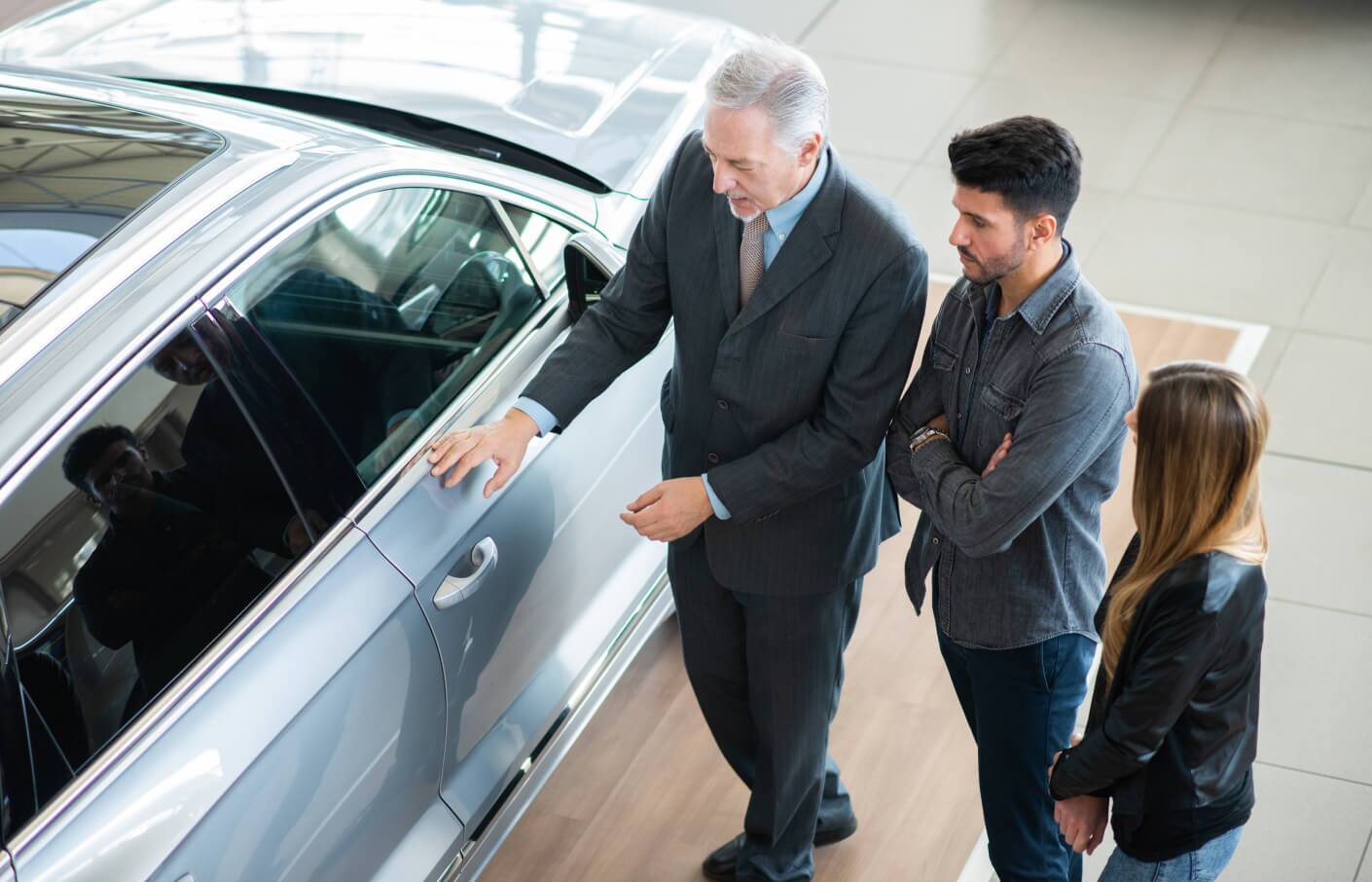High-angle side view of a car dealer showing a silver sedan to a couple in a showroom.