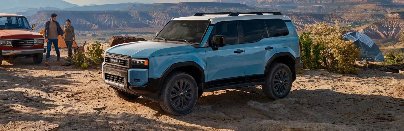 front quarter view of the 2024 Toyota Land Cruiser with a couple and their dog in the background against the backdrop of mountains