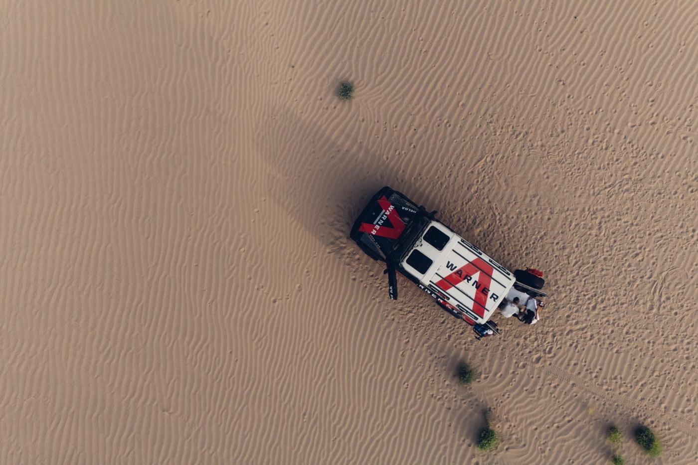 Overhead drone view of Helga parked in rippled sand, the Warner roof graphic sharp, the crew working at the open tailgate.