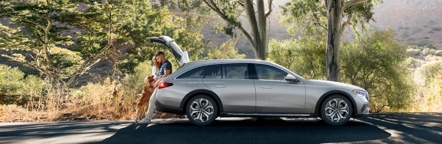 side view of the 2025 Mercedes-Benz E-Class All-Terrain Wagon with a couple and a dog standing behind it