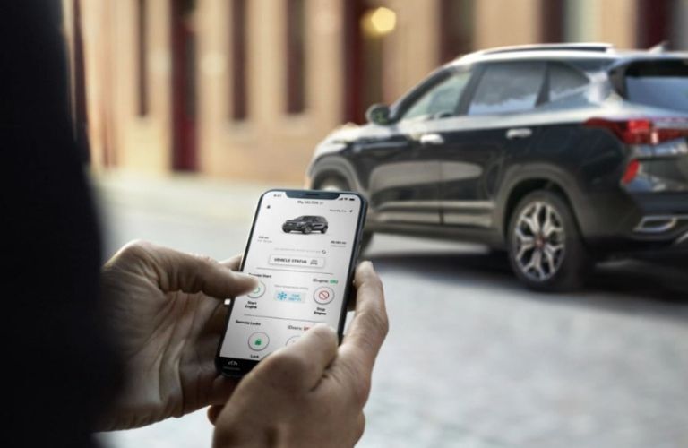 A female hand holding a smartphone with UVO link app while the Seltos is parked in the background