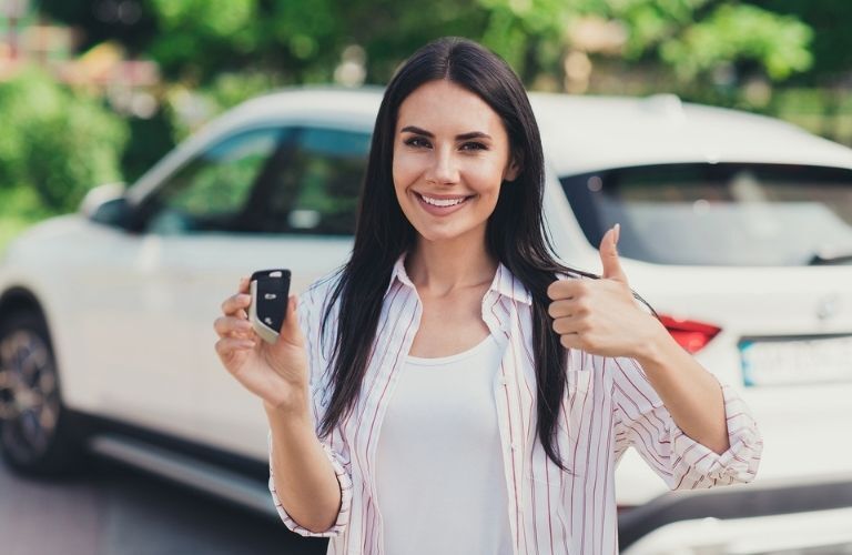 A woman holding a car key and showing thumbs up gesture with a blurry white car in the background