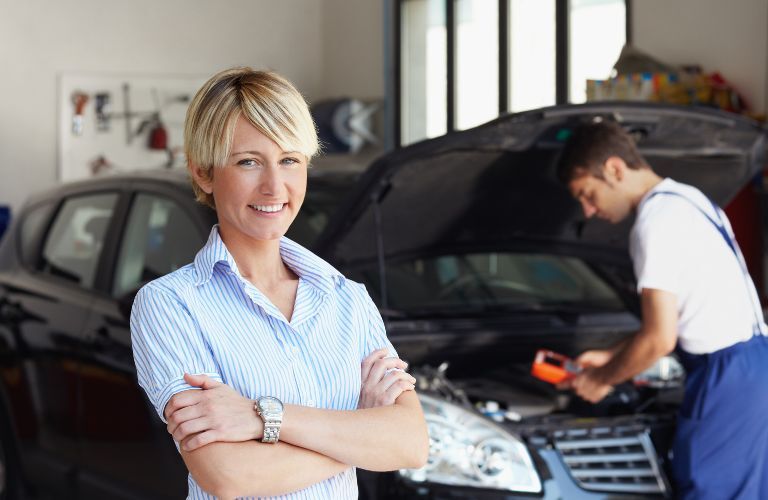 A smiling woman posing in front of a mechanic working on a vehicle at a service center