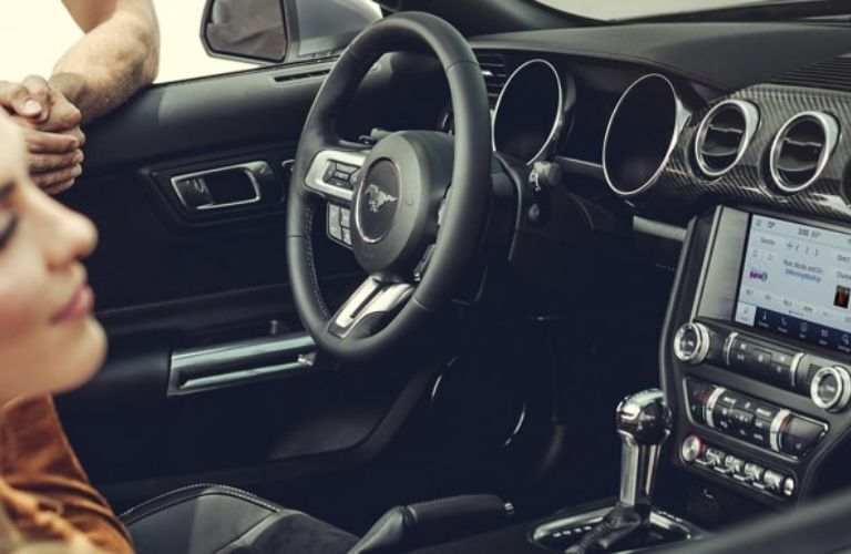 Close up of a steering wheel area of a Ford Mustang with a lady sitting inside is shown.