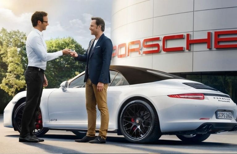 Image showing a car salesman handing over a key to a man standing next to a Porsche sports car outside a Porsche dealership
