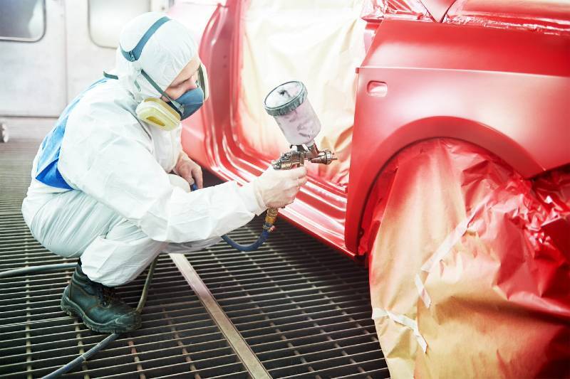 A professional car painter in respirator and body suit painting a red car
