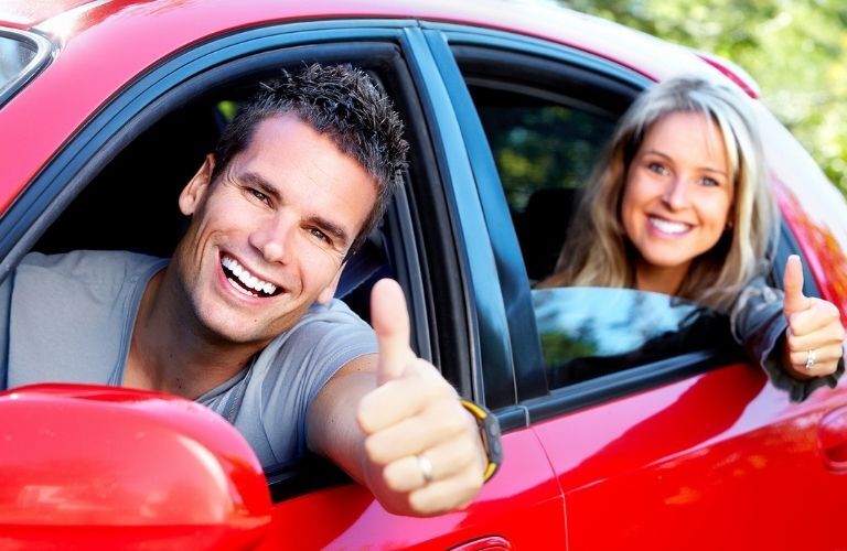 A happy couple in a red car doing the thumbs up gesture