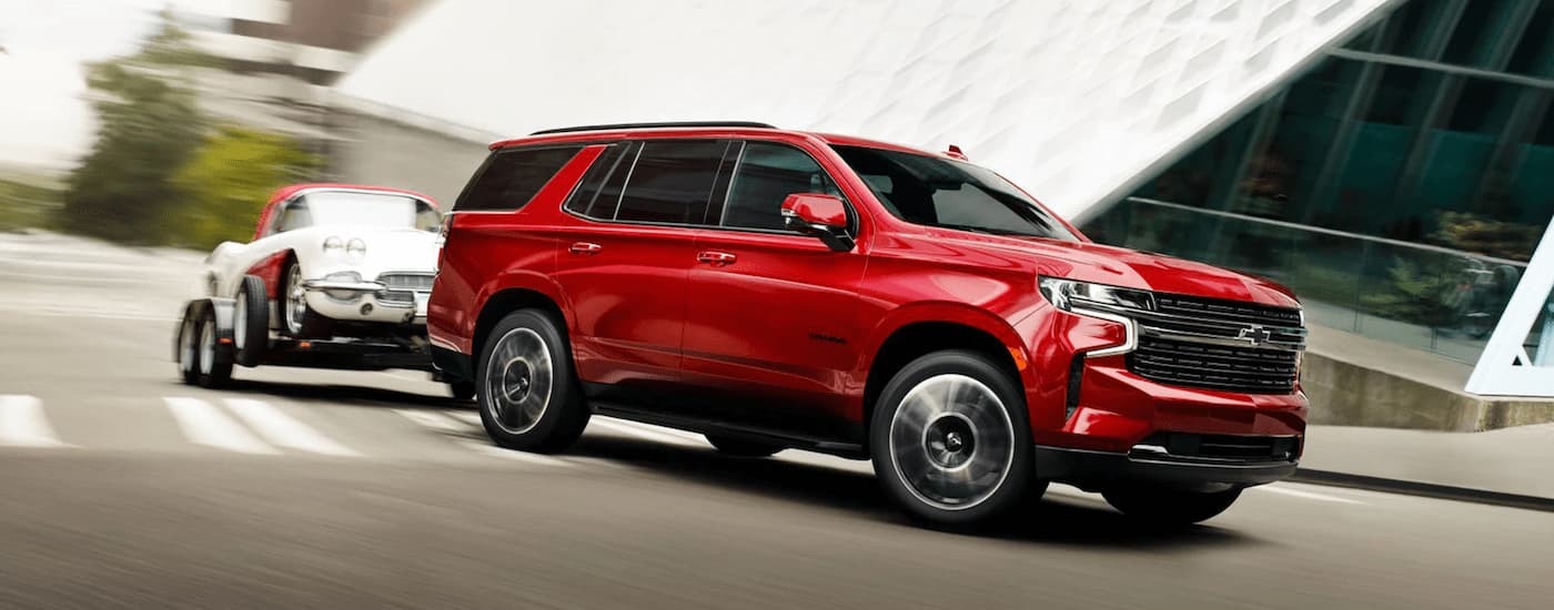 A red 2023 Chevy Tahoe is towing a white car on a trailer on a city street.