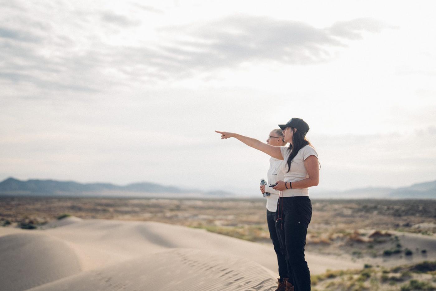 Racine Warner and Diane Walsh standing on a dune crest in early light, one pointing across the flats.