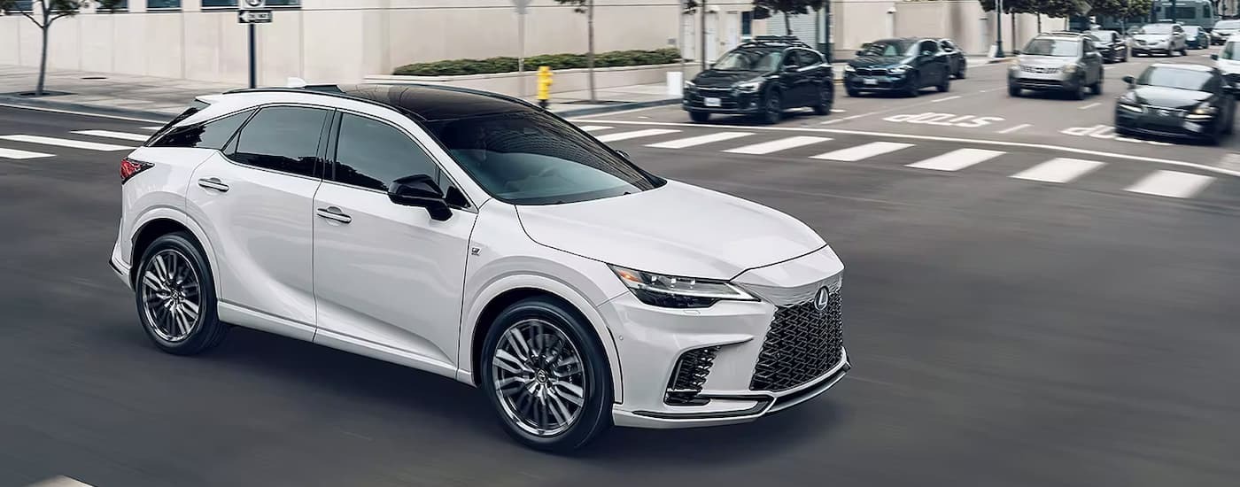 A white 2023 Lexus RX is driving through an intersection near a used car dealership in Jersey City.
