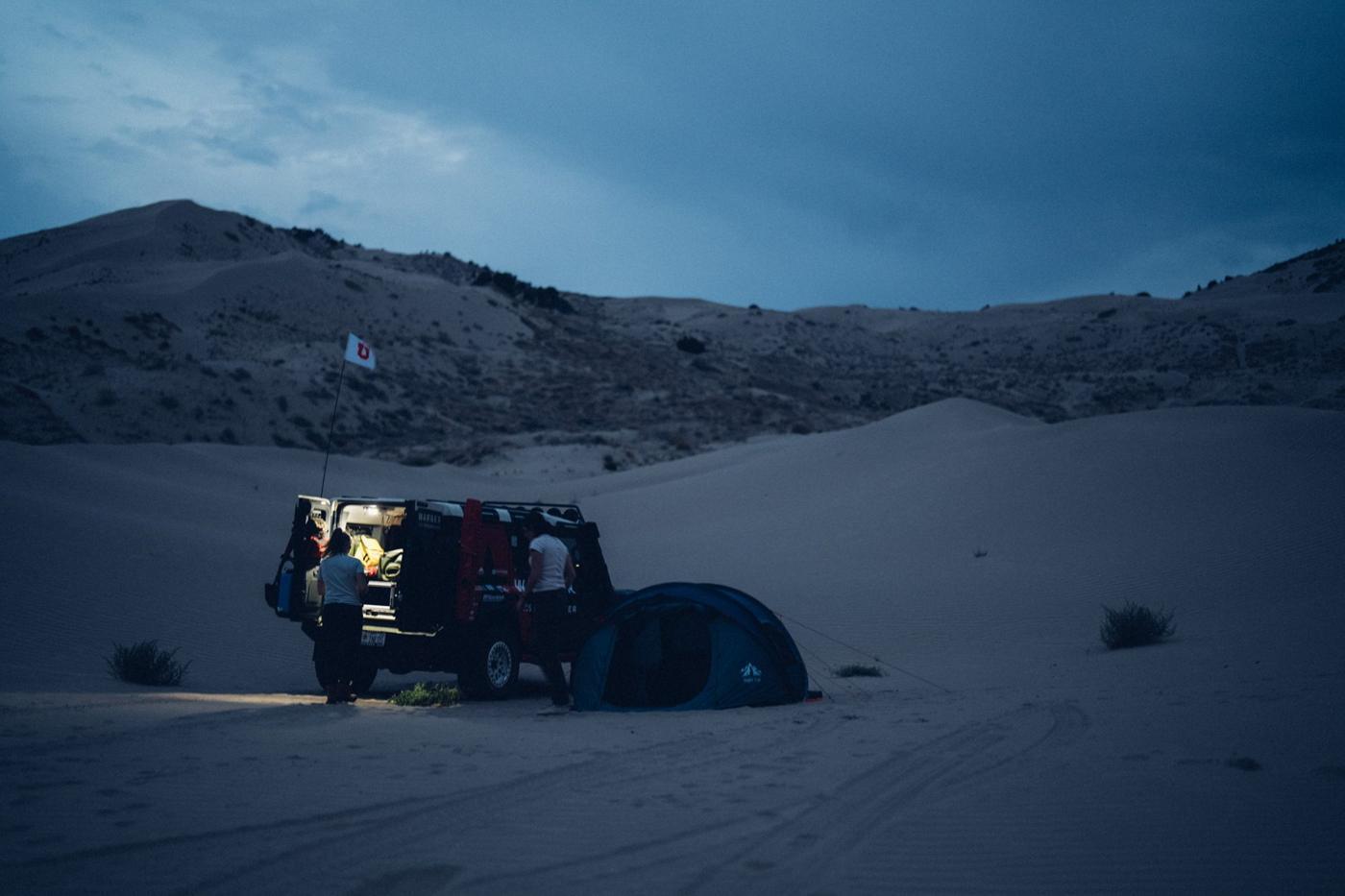 Helga at dusk with a tent pitched beside her, the crew packing the tailgate by cargo light under darkening dunes.