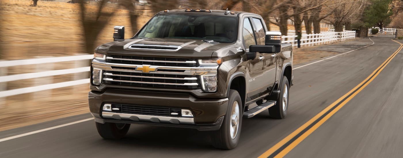 Brown 2023 Chevy Silverado 2500HD driving on a white fence-lined road