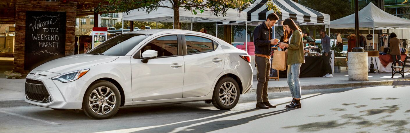 Two people standing next to white 2019 Toyota Yaris parked near marketplace