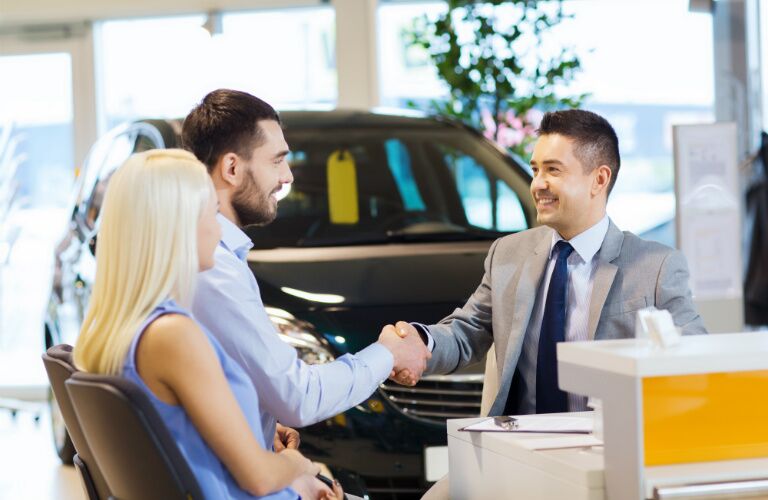 man and woman sitting at desk with salesman in car dealership and shaking hands