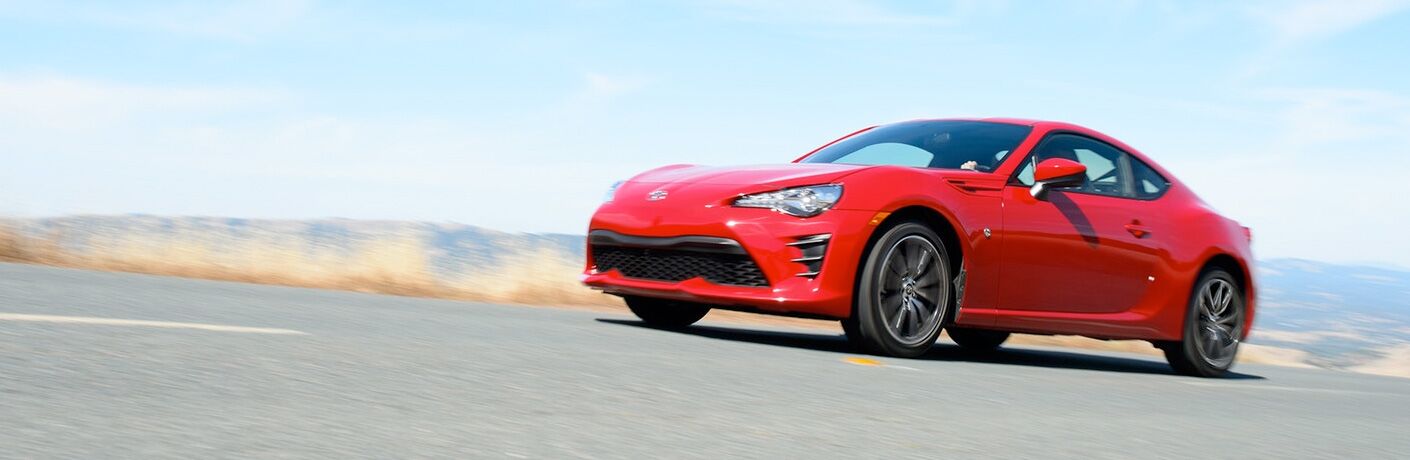 Exterior view of a red 2018 Toyota 86 driving down a country highway with mountains in the far background