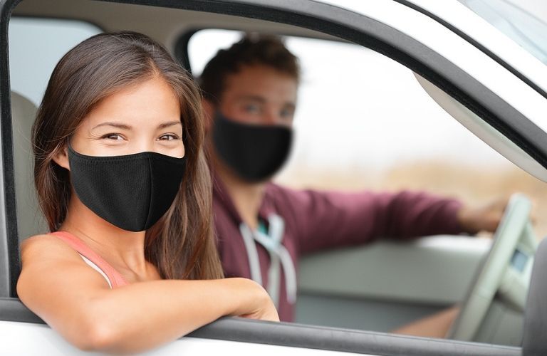 Imagse showing a young woman wearing a mask seated in a car with a blurred out young man at the steering wheel