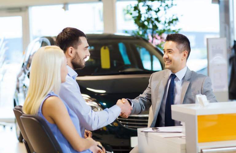 car dealership employee shaking hands with seated customers with a car in the background